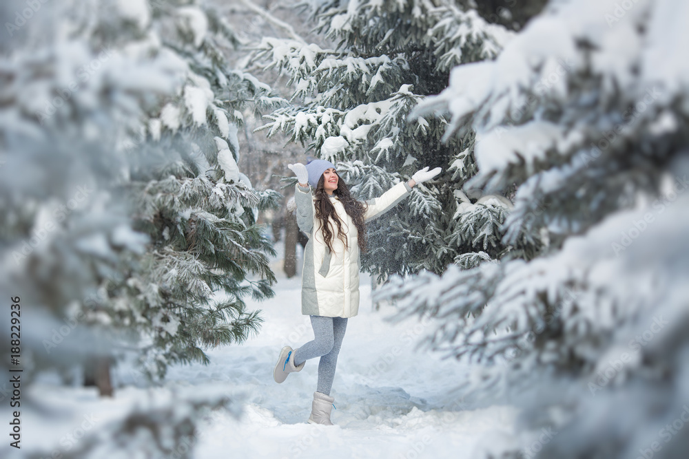 Smiling young woman in a white-grey clothes plays between a firs in the winter
