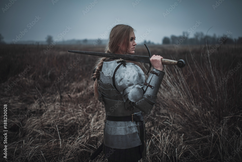 Fototapeta premium Girl in image of Jeanne d'Arc in armor and with sword in her hands stands on meadow. Back view.