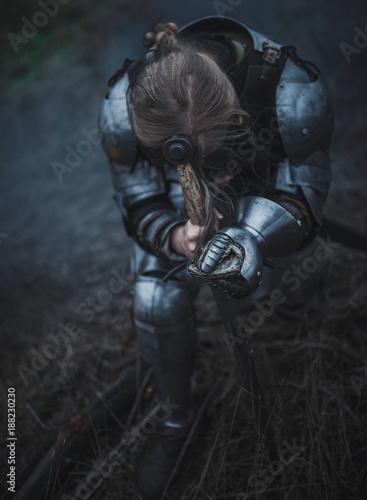 Fotografie Girl in image of Jeanne d'Arc in armor and with sword in her hands kneels against background of dry grass