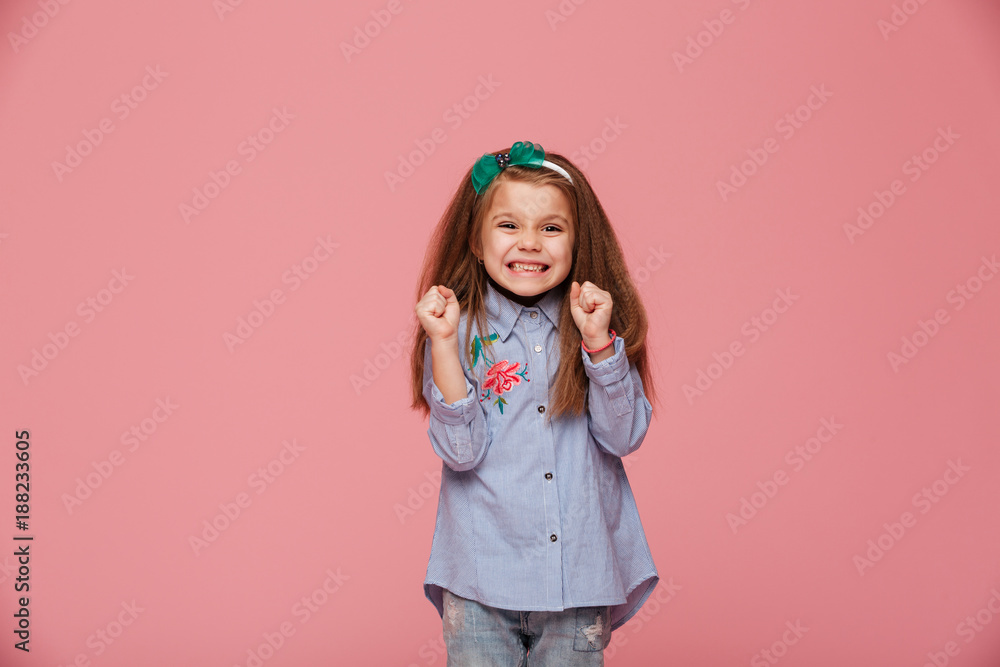 Smiling girl model in hair hoop and fashion clothes expressing happiness gesturing with clenched fists against pink background Smiling girl model in hair hoop and fashion clothes expressing happiness gesturing with clenched fists against pink background