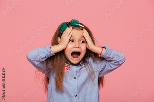 Thrilled little girl reacting emotionally grabbing head with both hands being shocked screaming over pink background