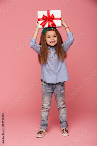 Full-length portrait of joyous female kid holding gift box with red ribbon bow on her head feeling pleasure over pink background