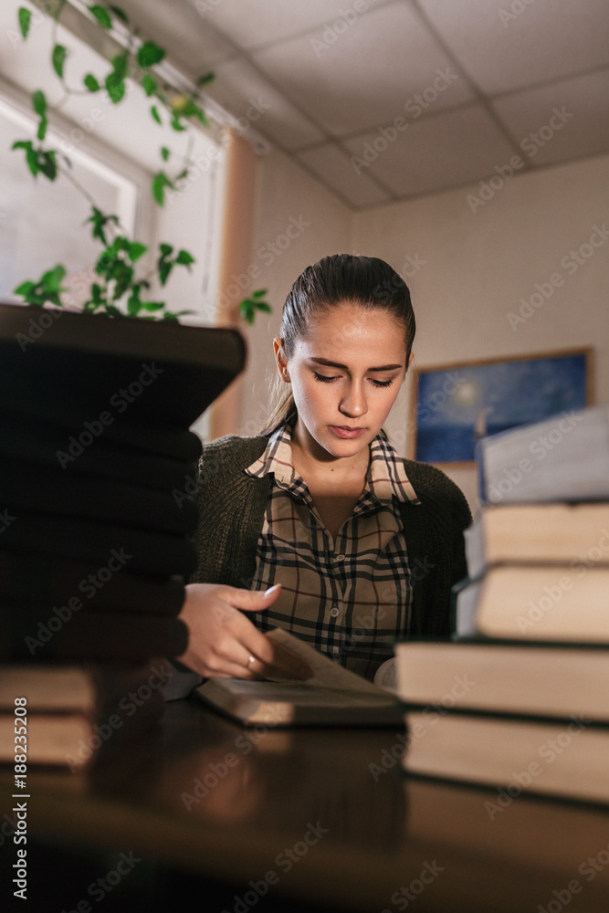 a beautiful slim girl in a knitted green cardigan with a dark hair holds her hand near the chin and sees at right near piles of large books in the library