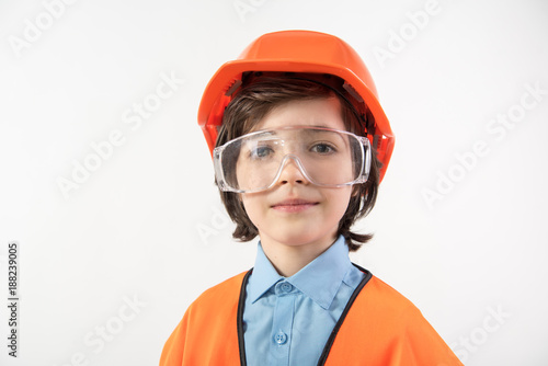 Portrait of dreamy kid preparing to become an architect. He is wearing orange helmet and safety glasses. Isolated on background © Yakobchuk Olena