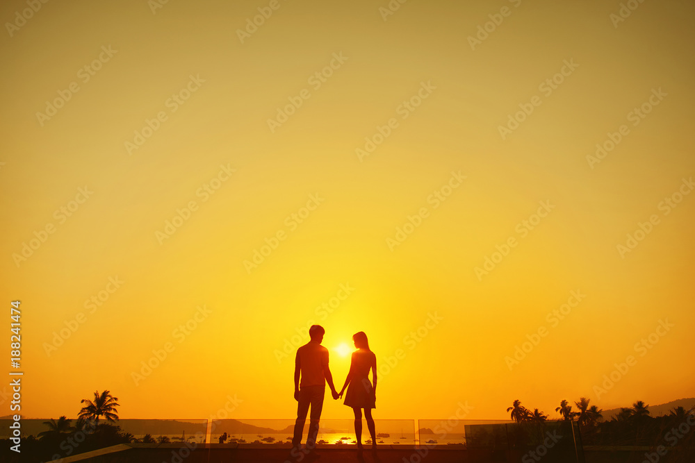 Silhouette of couple standing on the roof of building with view on bay and palms at sunset.