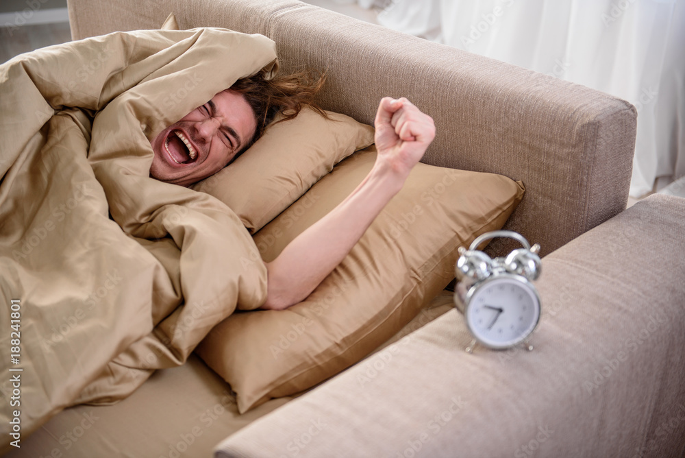 Grumpy man waving his hand at the clock and crying in bedroom. He is lying in bed with a grimace on his face Grumpy man waving his hand at the clock and crying in bedroom. He is lying in bed with a grimace on his face