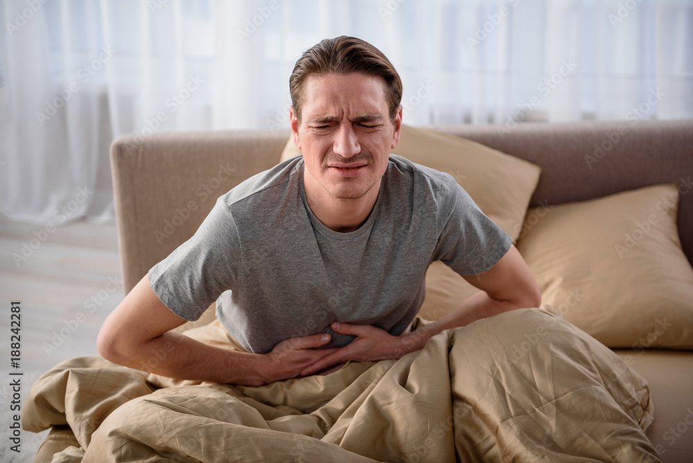 Waist up portrait of sick man sitting on bed with closed eyes. He is terribly ill and having a stomachache Waist up portrait of sick man sitting on bed with closed eyes. He is terribly ill and having a stomachache