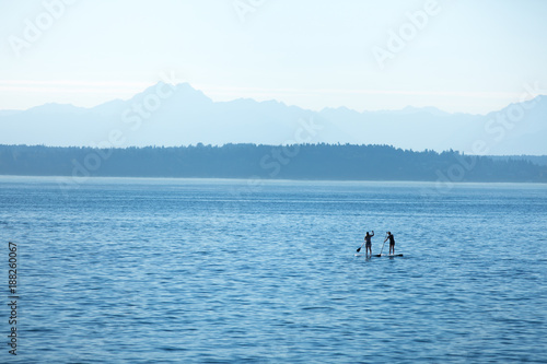 Two silhouetted stand up paddle board riders on a calm and blue ocean, with mountains on the horizon