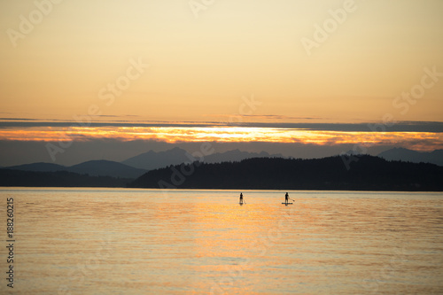 Two silhouetted stand up paddle board riders on a calm ocean, with mountains on the horizon and orange sunset