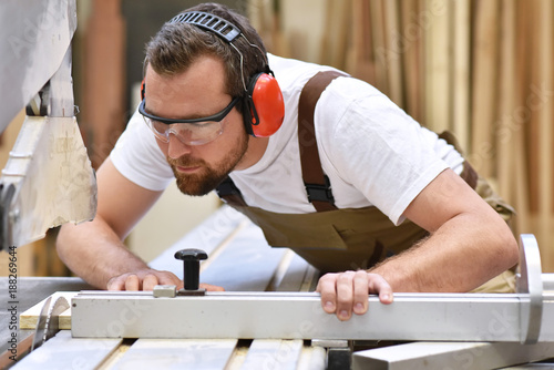 Tischler in einer Schreinerei sägt ein Holzbrett an einer Sägemaschine - Handwerker in Berufsbekleidung // Joiner saws a wooden board on a sawing machine - craftsmen in work clothing