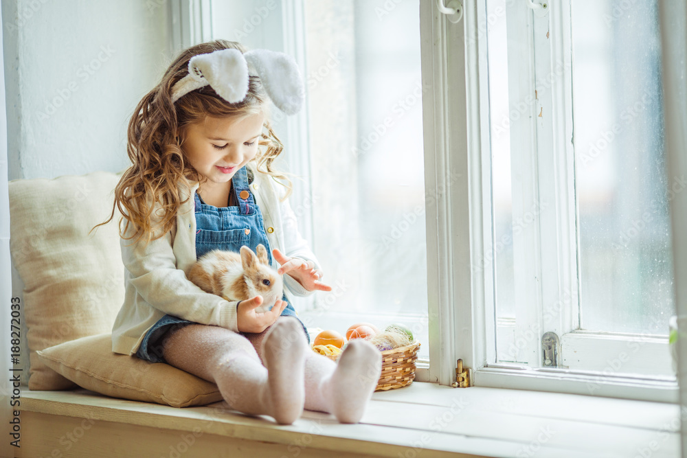 Fototapeta premium Cute happy little child girl is wearing bunny ears on Easter day holding little rabbit