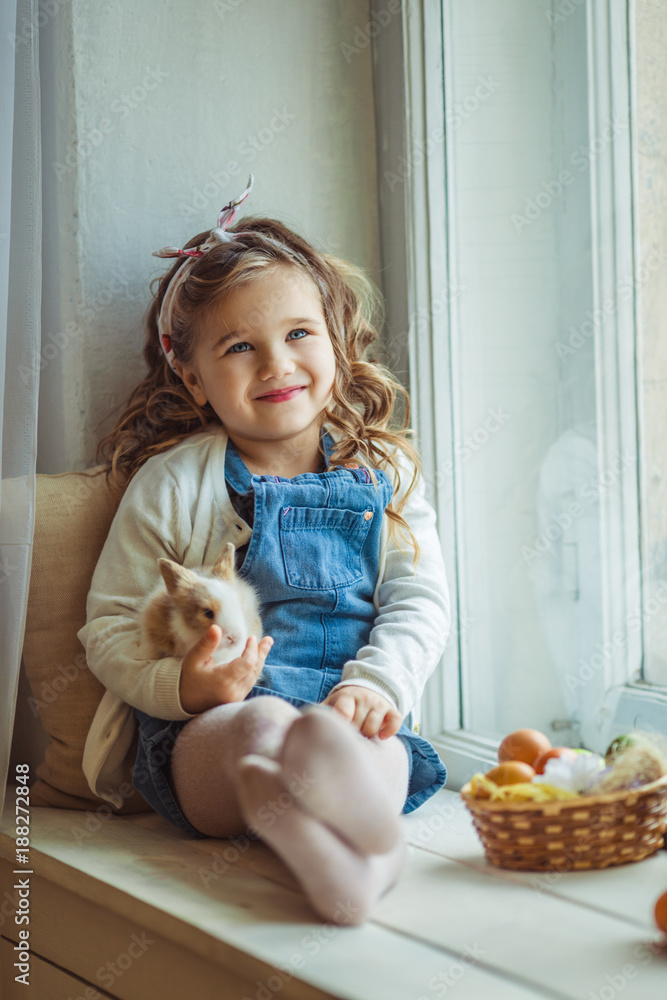 Obraz premium Beautiful happy child girl is sitting on window sill and holding her friend little colorful rabbit, Easter holiday concept