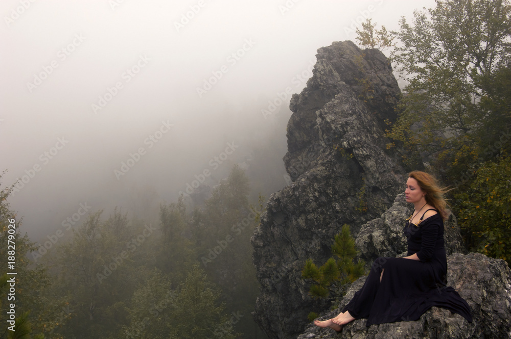 the redhead bare-footed woman in a black dress sits on a rock above the forest (the rock is the peak of Mount Kolpaki and the conditional border between Europe and Asia in the Middle Ural).. the redhead bare-footed woman in a black dress sits on a rock above the forest (the rock is the peak of Mount Kolpaki and the conditional border between Europe and Asia in the Middle Ural)..