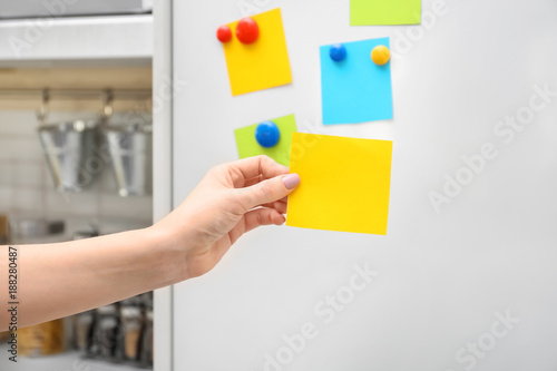 Woman holding empty note near refrigerator door in kitchen