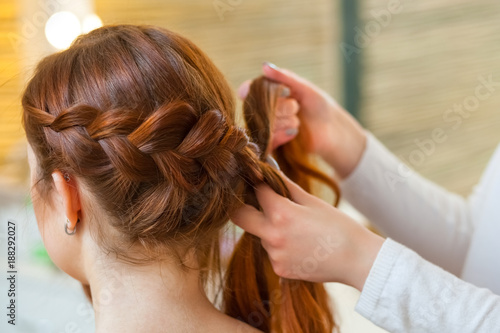 Beautiful girl with red hair, hairdresser weaves a braid close-up, in a beauty salon. Professional hair care and creating hairstyles.