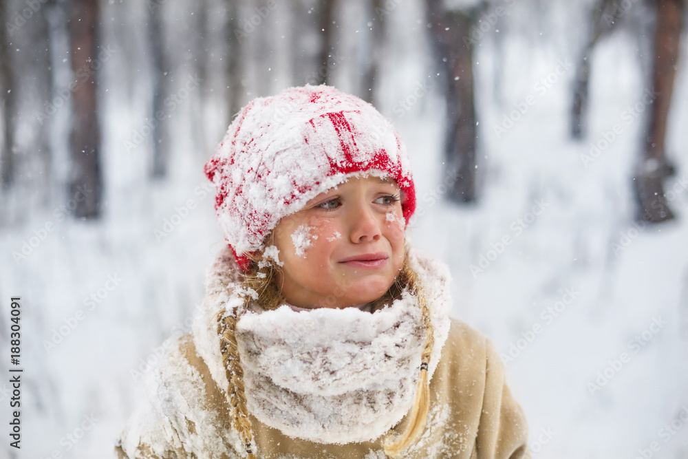 Crying girl wearing in red knitted hat in snowy winter park. Little sad child play with snow in forest. Family winter vacation with  kid. Young girl froze and cry