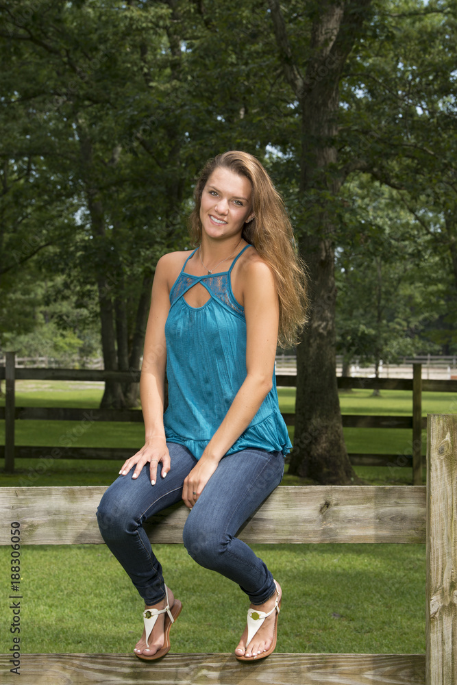 Obraz premium Beautiful teen girl poses while sitting on a wooden fence on farm in jeans and blouse