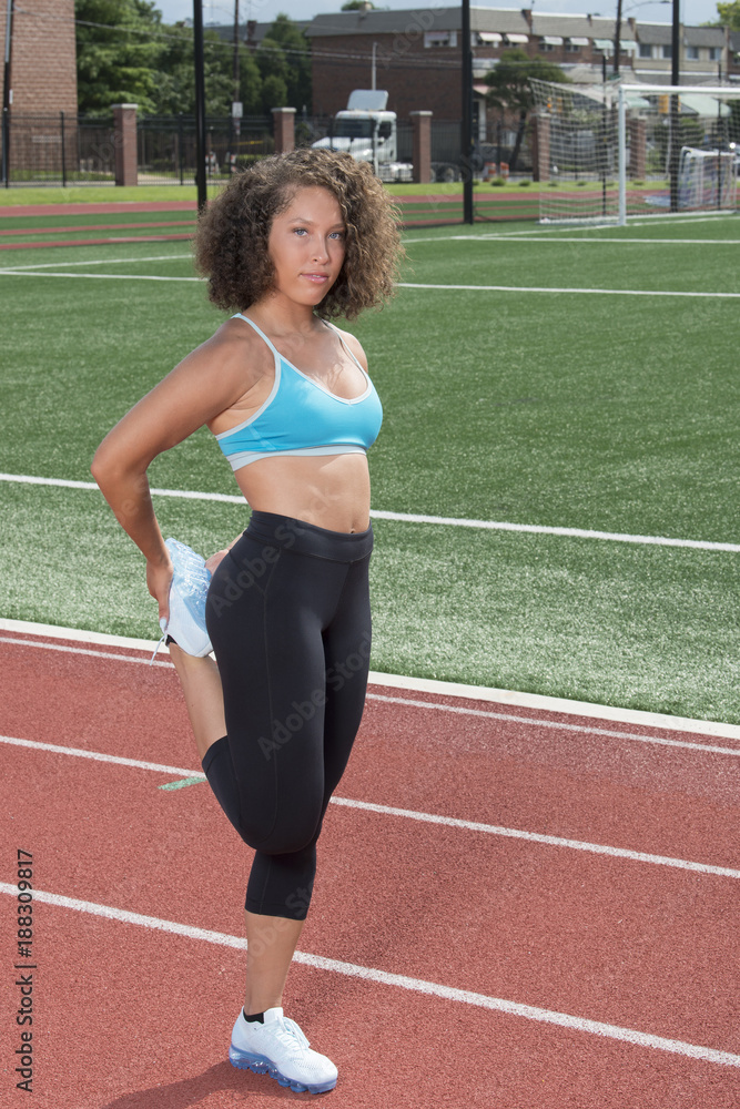 Obraz premium Beautiful young African-American fitness model stretches her leg on an outdoor track ready to work-out in blue sports bra and black pants