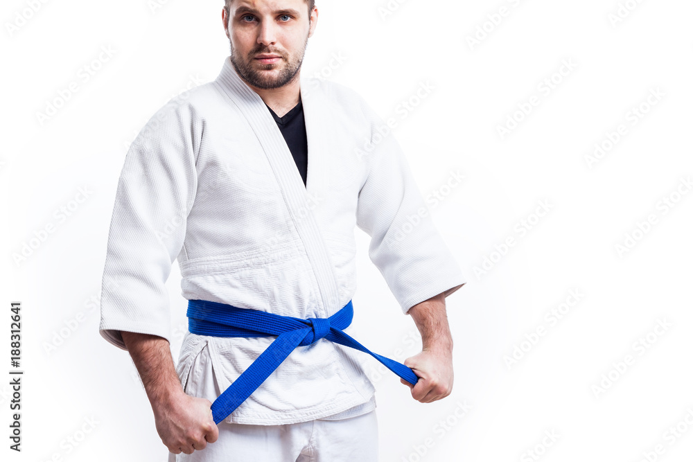 Young sporty man with dark hair in a white kimono with blue sambo belt, jiu  jitsu, judo stands and keeps his hands on a white isolated background,  front view Stock Photo