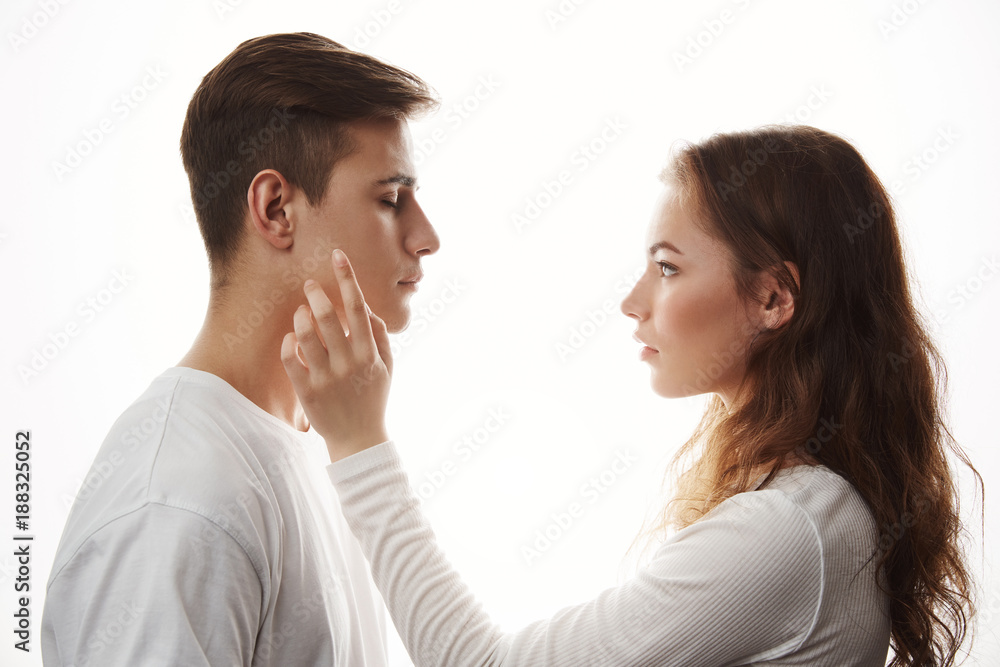 Sideways portrait of young couple in a white room. Girlfriend touches his boyfriends cheek showing love and care to him. She thinks if their future son will look like him Sideways portrait of young couple in a white room. Girlfriend touches his boyfriends cheek showing love and care to him. She thinks if their future son will look like him