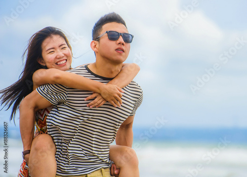  beautiful Asian Chinese couple with boyfriend carrying woman on her back and shoulders at the beach smiling happy in love enjoying holidays