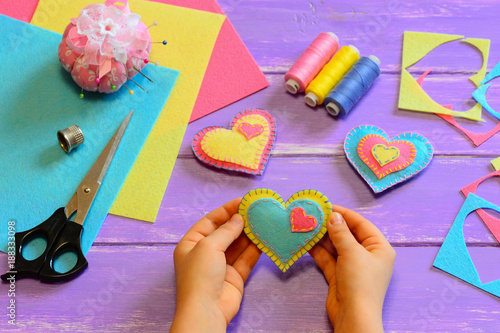 Child holds a felt heart gift in his hands. Child show a felt heart gift. Heart ornaments, scissors, felt sheets on a wooden table. Homemade Valentines Day decor crafts. Hand sewing projects for kids
