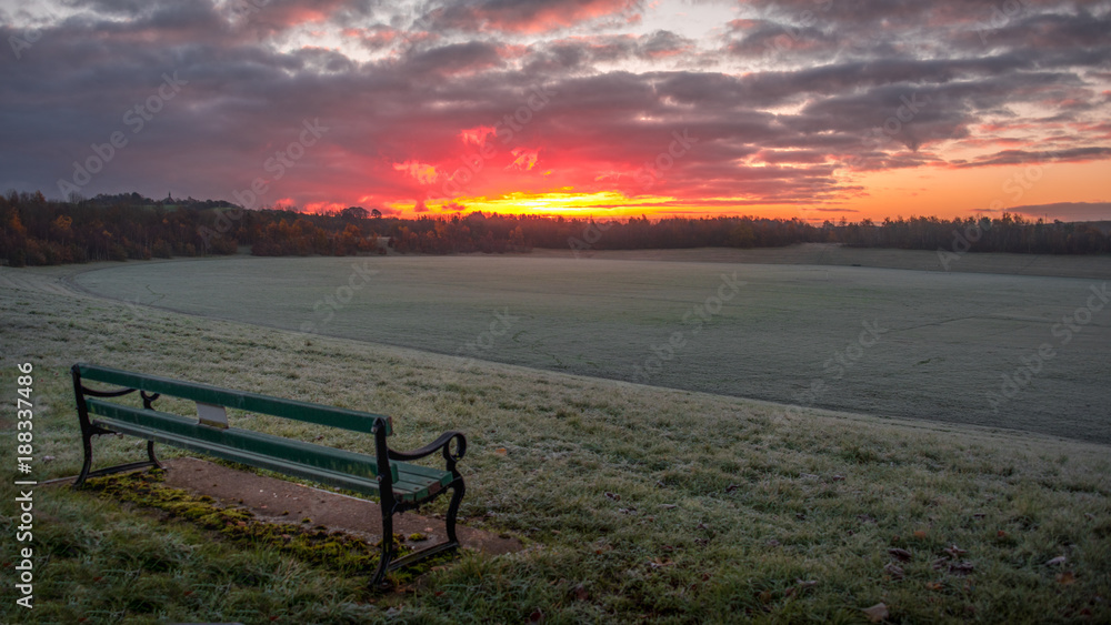 A beautiful early morning sunrise over an open frosty field parkland with an empty bench seat in the foreground