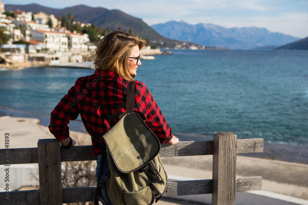 Woman in a shirt with a backpack is standing on a wooden pier and looking at the horizon. Sea coast, mountains. Woman is a traveler.