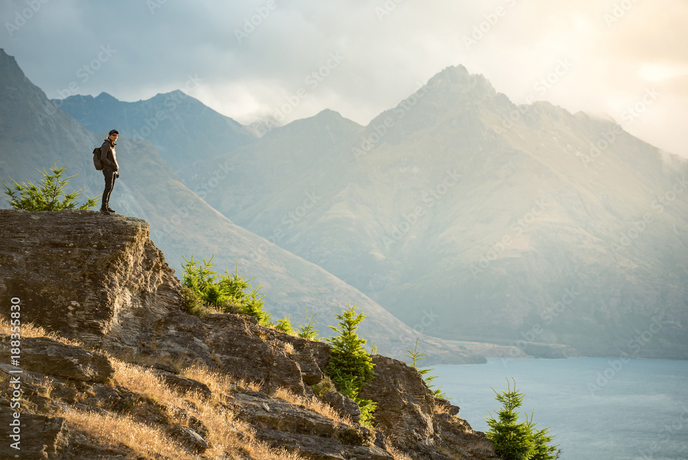Young Asian male traveler standing on the edge of vista lookout with mountain scenery and Lake Wakatipu in background, Watching sunset in Queenstown, South Island, New Zealand
