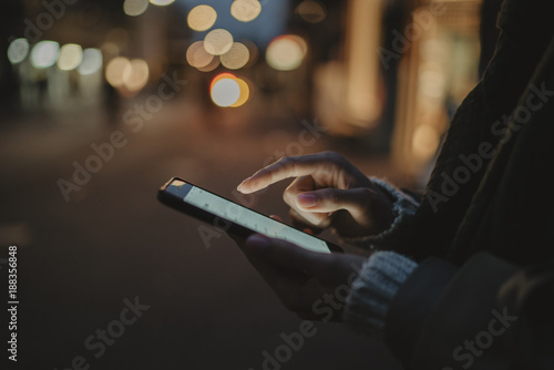 Cropped image of female hands using modern smartphone at night, bokeh light on the background