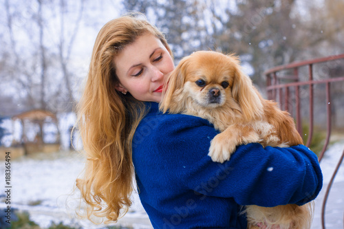 Woman pet owner with blue coat and golden hair walking during winter with her dog. Young lady with pekingese dog walking on the snowy field. Tender feeling with a dog 