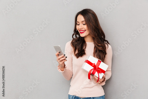 Portrait of attractive brunette female 30s winning silver new smartphone in lottery being joyful over gray wall © Drobot Dean