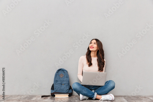 Horizontal image of female looking upward while reading or communicating online using silver laptop sitting with legs crossed on the floor over gray wall photo