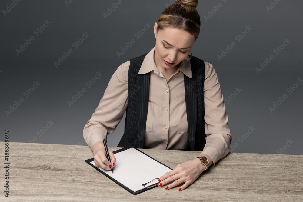 attractive kazakh businesswoman writing on clipboard at table isolated on grey attractive kazakh businesswoman writing on clipboard at table isolated on grey
