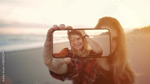 Cinemagraph of young teenage girl or student woman with blond hair and in red pleaded shirt smile and laugh, makes selfies during sunset at beach on summer vacation