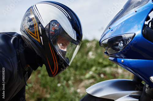 Fotografie Close up sideways view of blue motorcycle and self determined young male rider wearing black leather jacket and stylish protective helmet getting ready for race