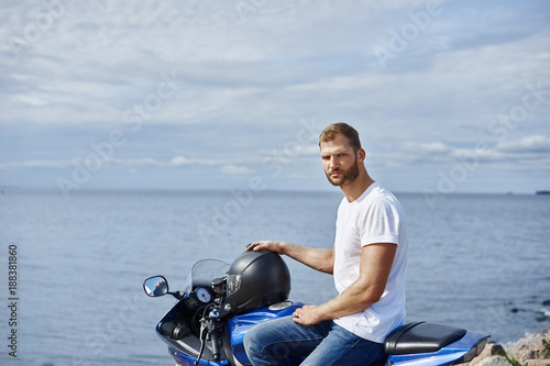 Attractive bearded young motorcyclist enjoying seaview while traveling on his motorcycle along seashore, posing against blue sky and sea background with copy space for your advertising content