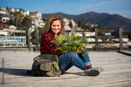 Happy girl traveler sits on a wooden pier and holds a bouquet of flowers of mimosa in hands. Next to the woman is a backpack. Sea and mountains on the horizon. Professional lighting.