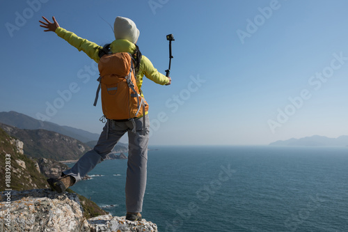 Young woman hiker using action camera mounted on selfiestick taking selfie at seaside