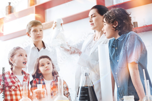 Can you see the reaction. Amazed up and coming chemists looking at a chemical reaction in a fuming filter flask held by a teacher of chemistry wearing a laboratory coat.