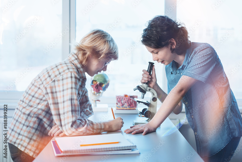 Obraz premium Noting the details. Intelligent attentive inspired boy telling his friend what he observing through the microspore while fulfilling practical assignment in biology class