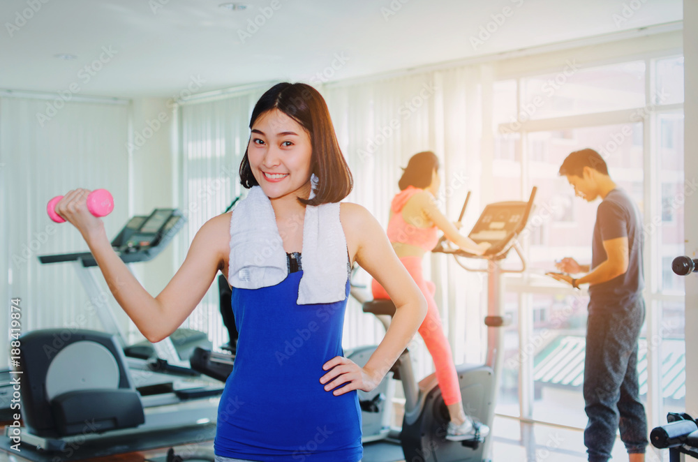 young asian pretty woman doing exercises with dumbbell at biceps in fitness gym with people background, bodybuilder, healthy lifestyle, exercise fitness, workout and sport training concept, soft focus young asian pretty woman doing exercises with dumbbell at biceps in fitness gym with people background, bodybuilder, healthy lifestyle, exercise fitness, workout and sport training concept, soft focus