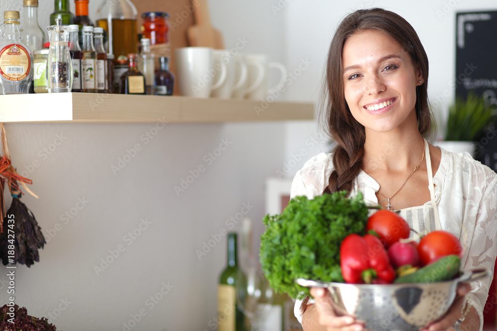 Smiling young woman holding vegetables standing in kitchen. Smiling young woman Smiling young woman holding vegetables standing in kitchen. Smiling young woman