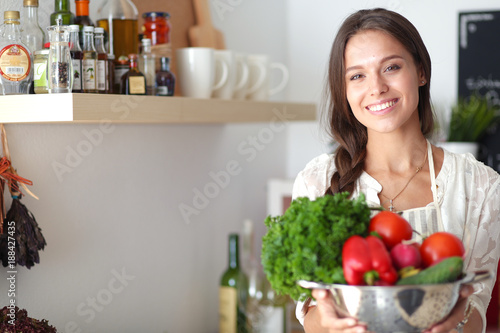Smiling young woman holding vegetables standing in kitchen. Smiling young woman