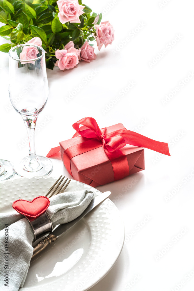 Valentines day dinner with table place setting with red gift, glass for champagne, heart ornaments with silverware on white. Close up. Valentine's card.