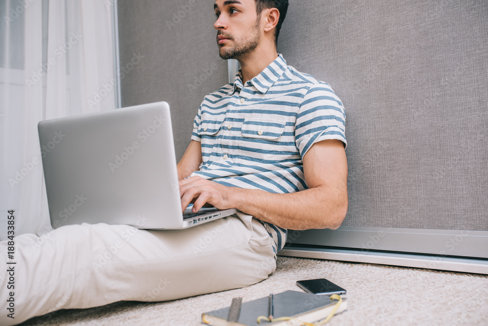 Cropped portrait of cheerful young Caucasian man in stylish shirt sitting on the floor at home with open laptop pc on his lap, notice on paper for business startup. People and lifestyle concept.