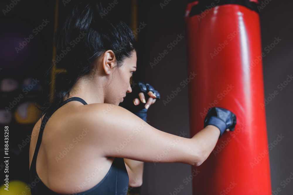 Sport, fitness, lifestyle, people and motivation concept. Rear view of athlete brunette female with red towel on neck punching the red bag in kickboxing gloves at the gym. Woman boxer workout.