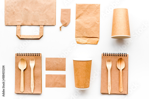 Disposable tableware. Paper cup, spoon, fork near brown paper bag, label and notebook top view mock up pattern white background