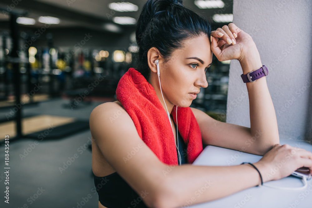 Side view of tired young beautful fitness woman leaning to sill in the gym after cardio. Sportswoman taking a break from her workout with red towel on neck and earphones listening the music. Side view of tired young beautful fitness woman leaning to sill in the gym after cardio. Sportswoman taking a break from her workout with red towel on neck and earphones listening the music.