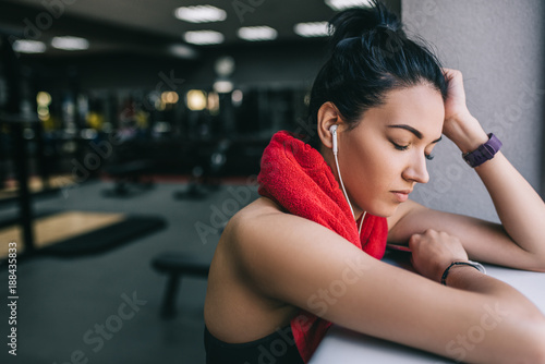 Portrait of young woman after her workout with closed eyes with earphones listening music with copyspace for text. Female athlete taking rest after fitness training at gym with red towel on neck.